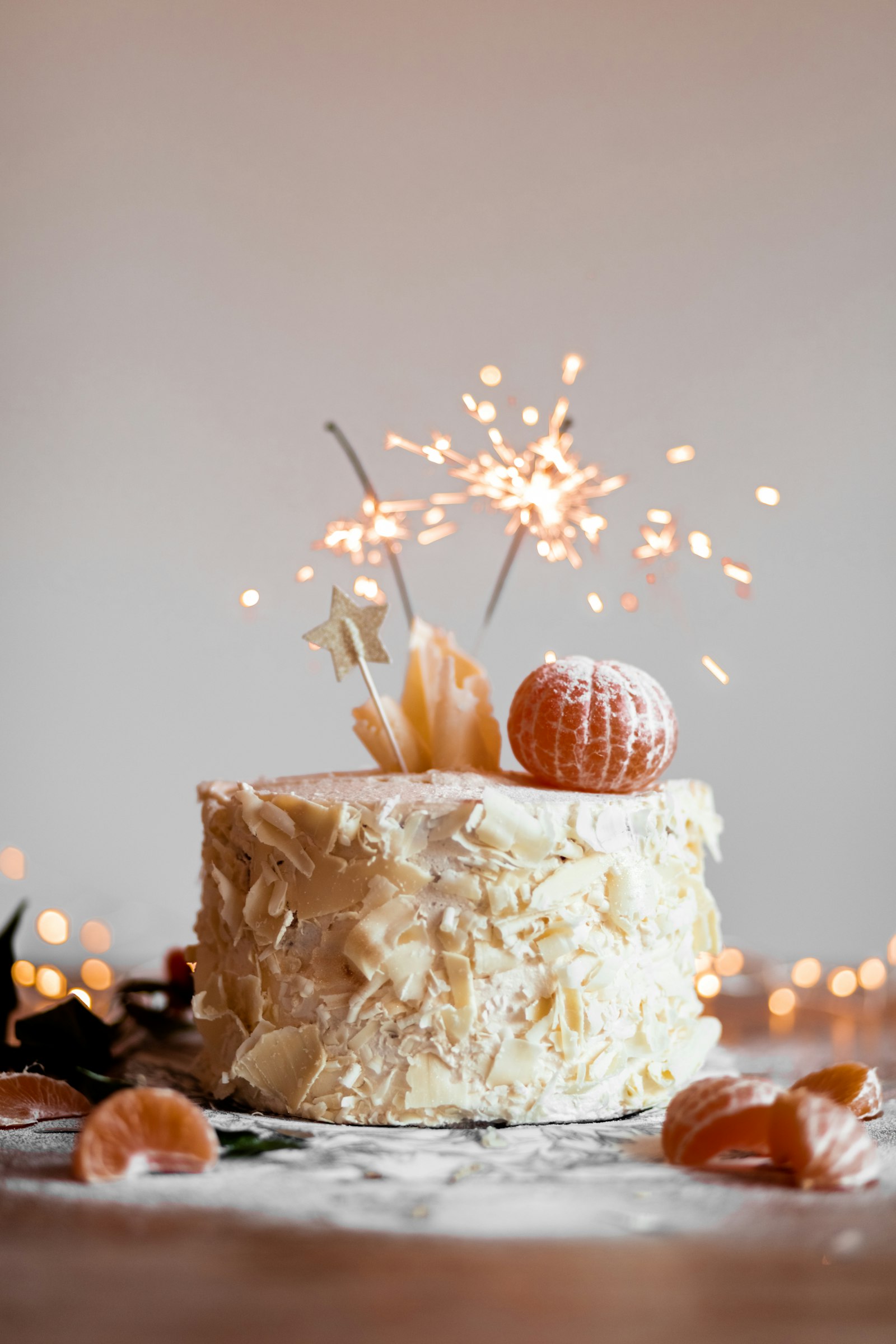 Pastel cubierto de virutas de chocolate blanco con luces de bengala encendidas y una mandarina. Imagen representativa.